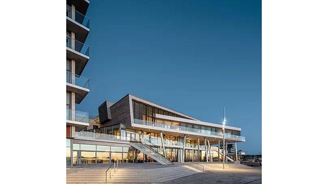 Modern byggnad med stora fönster och trappor, belägen vid vattnet under en klar kvällshimmel.