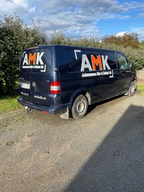 En svart skåpbil med texten "AMK" på sidan, parkerad på en grusväg omgiven av grönska.