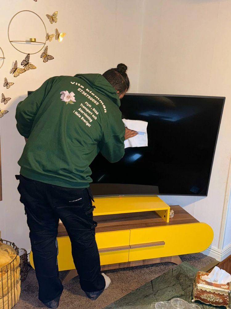 En person i grön jacka rengör en stor svart TV på en gul TV-bänk i ett vardagsrum.