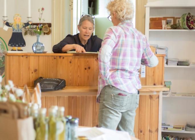 Kvinna i blus pratar med en annan kvinna vid en trädisk i en butik med varor i bakgrunden.