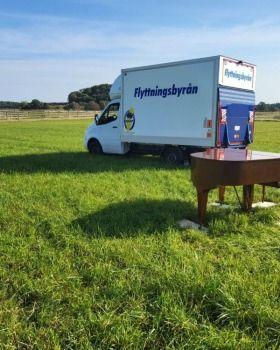 En flyttbil från Flyttningsbyrån står på en grön äng bredvid ett piano.