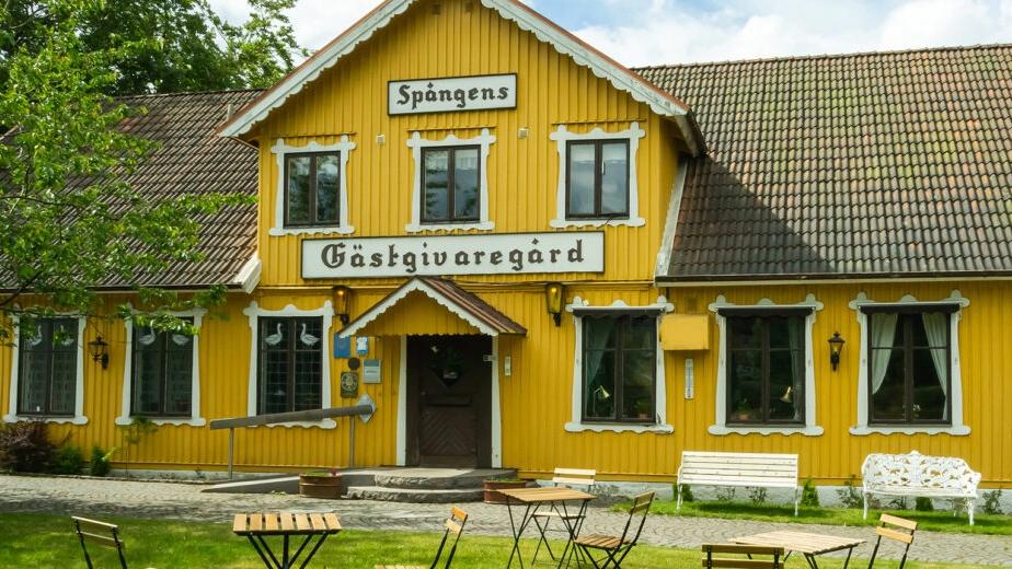 Gul gästgiveri med träfasad och bänkar i trädgården, omgiven av grönska och blå himmel.