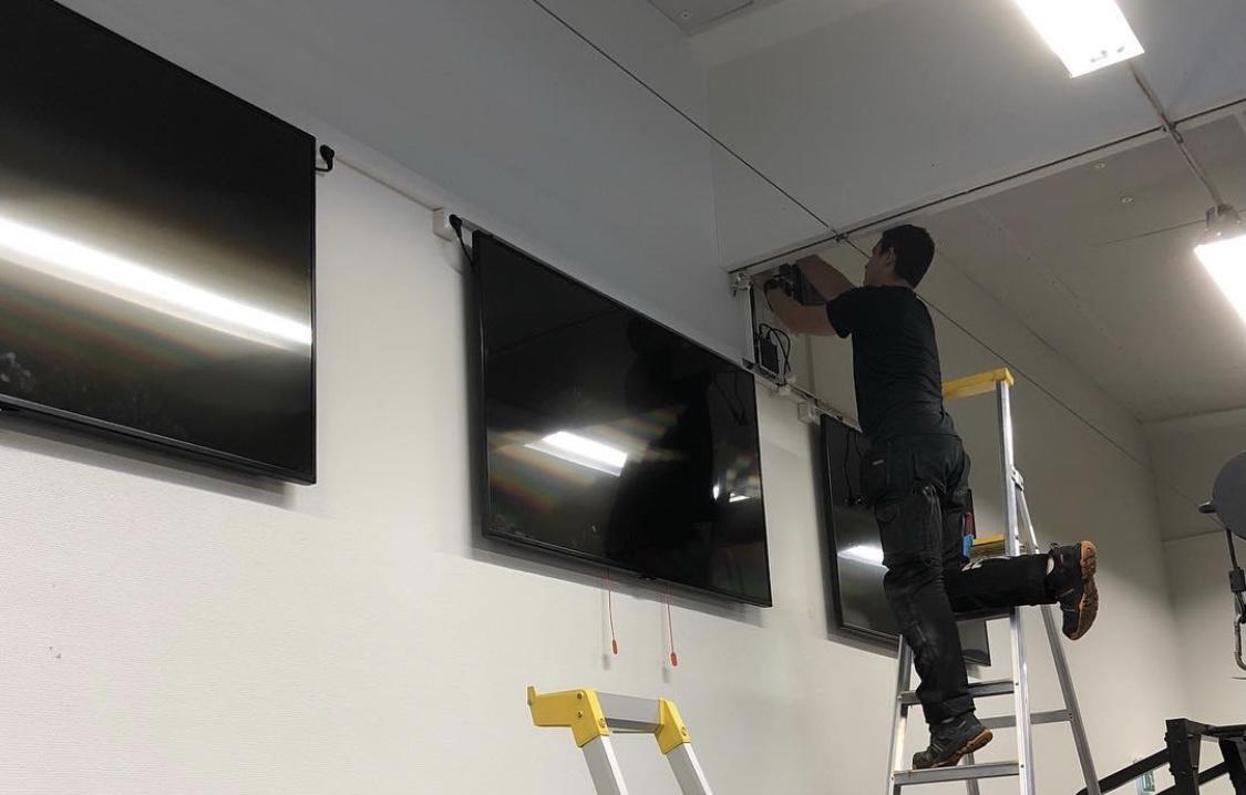 En tekniker installerar TV-apparater på en vägg med hjälp av en stege och verktyg.