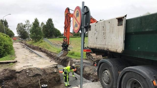 En grävmaskin arbetar på en väg med en skylt som visar att parkering är förbjuden, medan en arbetare övervakar.