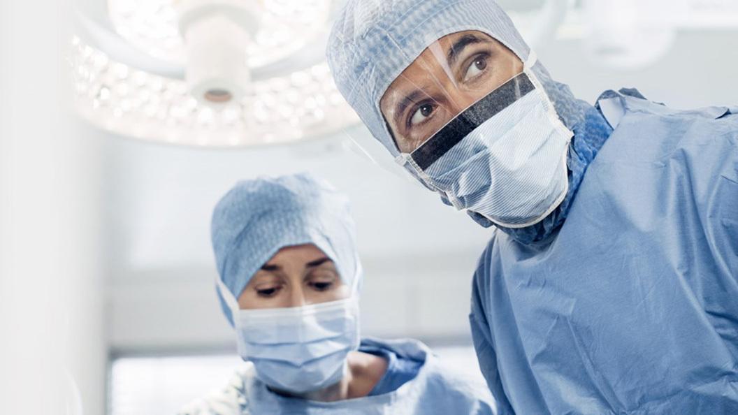 Två kirurger i operationskläder och munskydd, fokuserade på en patient under en operation.