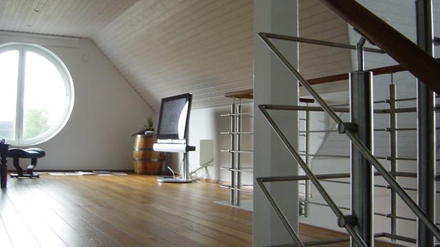 Ljust och rymligt loft med rund fönster, trappa i metall och trägolv. En tv står på en möbel i bakgrunden.