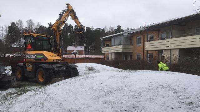 En grävmaskin arbetar på en snötäckt tomt framför en lägenhetsbyggnad. En arbetare syns i bakgrunden.