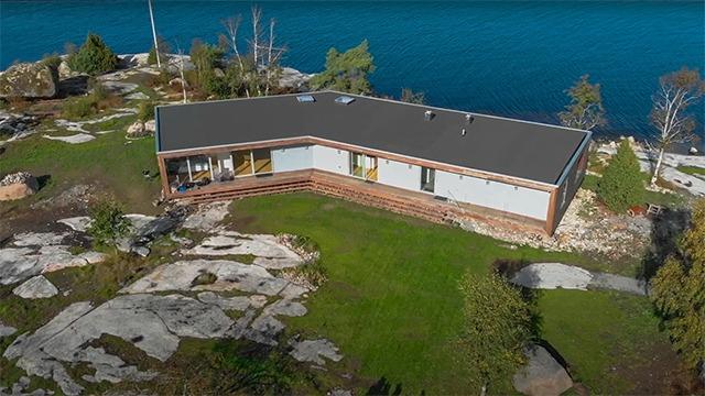 Ett modernt hus med stora fönster beläget vid vattnet, omgiven av klippor och grönska.