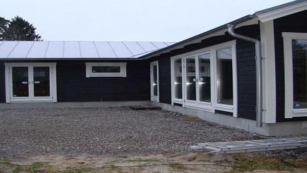 Nybyggd enplansvilla med mörk fasad och stora fönster, omgiven av grus och grönska.