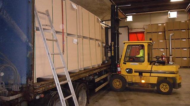 En gaffeltruck lastar stora kartonger i en lastbil i ett lager. Stora lådor syns i bakgrunden.