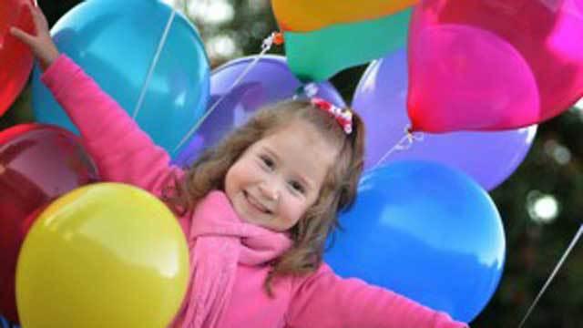 En glad flicka i rosa tröja håller upp färgglada ballonger och ler mot kameran.