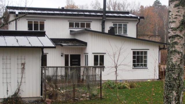 Ett vitt hus med svart tak och stora fönster, omgiven av en grön trädgård och träd.