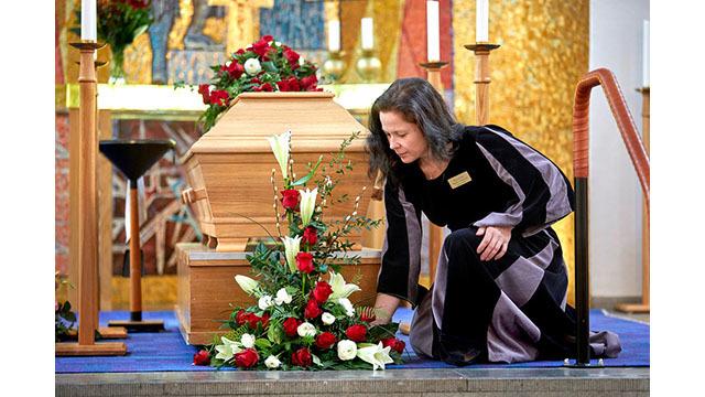 En kvinna i svart klänning placerar blommor vid en kista i en kyrka, omgiven av vackra dekorationer.