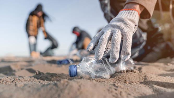 En hand i handskar plockar upp en plastflaska från sanden under en strandstädning. I bakgrunden syns fler personer som städar.