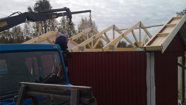 Byggarbetare som lyfter takstolar med en kran på en röd byggnad under konstruktion.