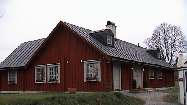 Ett rött hus med svart tak och vita fönster, omgiven av grön gräsmatta och träd.
