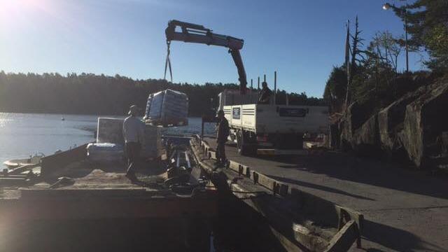 En kran lyfter en metallbehållare från en båt vid en kaj med skogiga öar i bakgrunden.