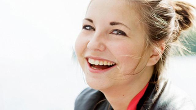 Leende kvinna med långt hår som sitter utomhus vid vattnet, klädd i en svart jacka.