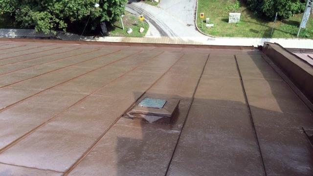 Tak med brunt takmaterial och en ventilationsöppning, omgiven av grönska och en väg i bakgrunden.