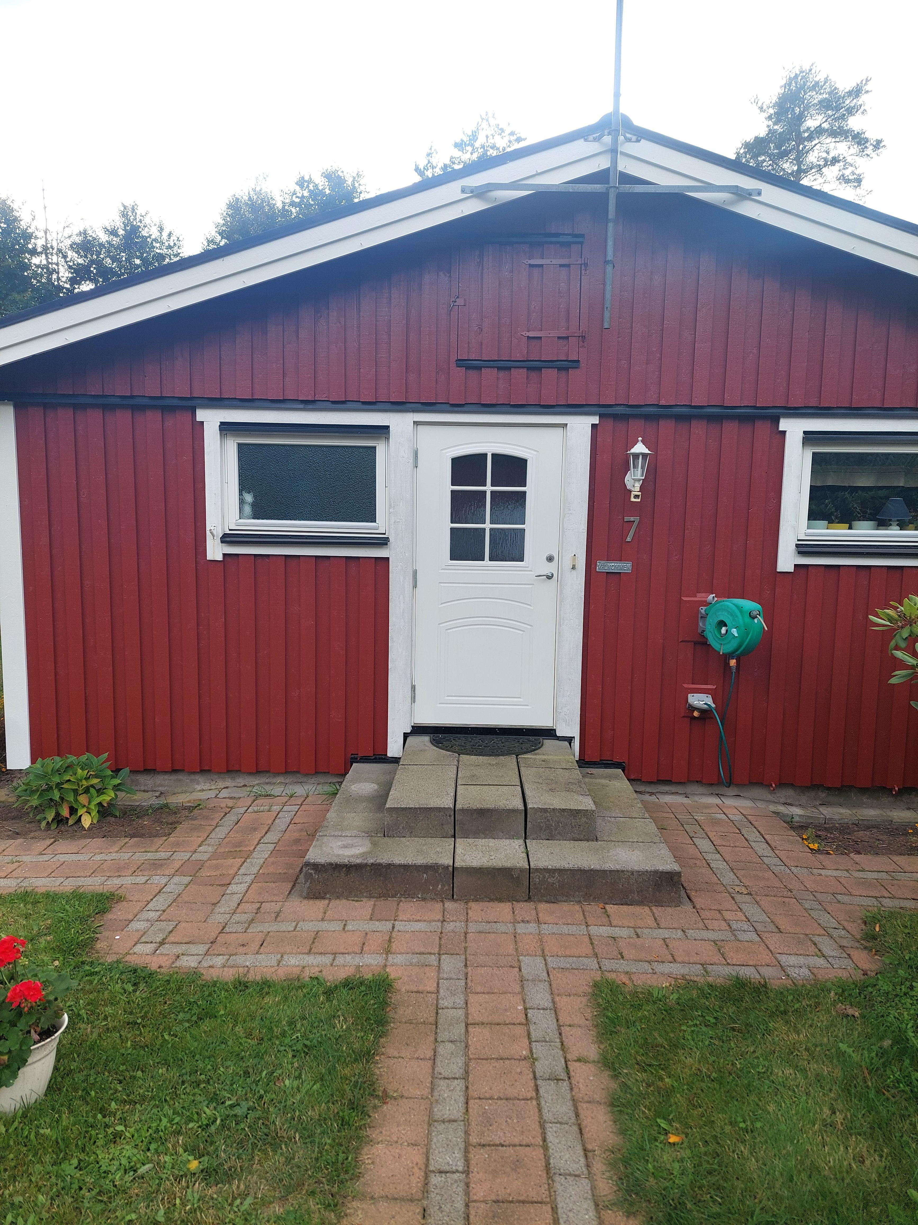 Röd stuga med vit dörr och stenlagd entré, omgiven av grön gräsmatta och blommor.