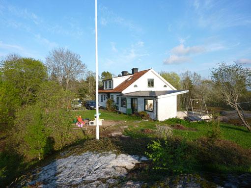 Ett vitt hus med röd takpanna omgiven av grönska och träd, beläget på en kulle under en blå himmel.