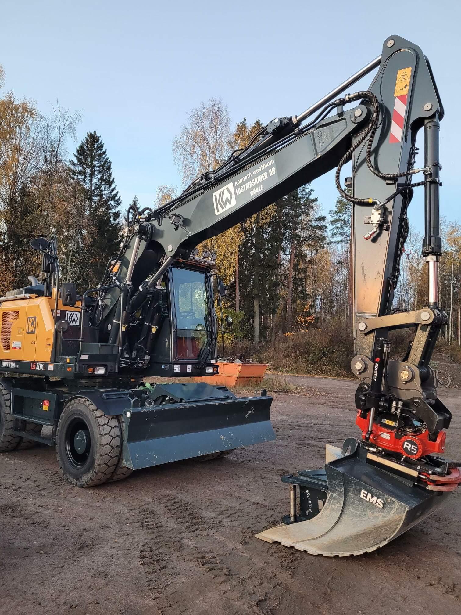 En grävmaskin med en skopa står på en byggarbetsplats omgiven av träd och höstfärger.