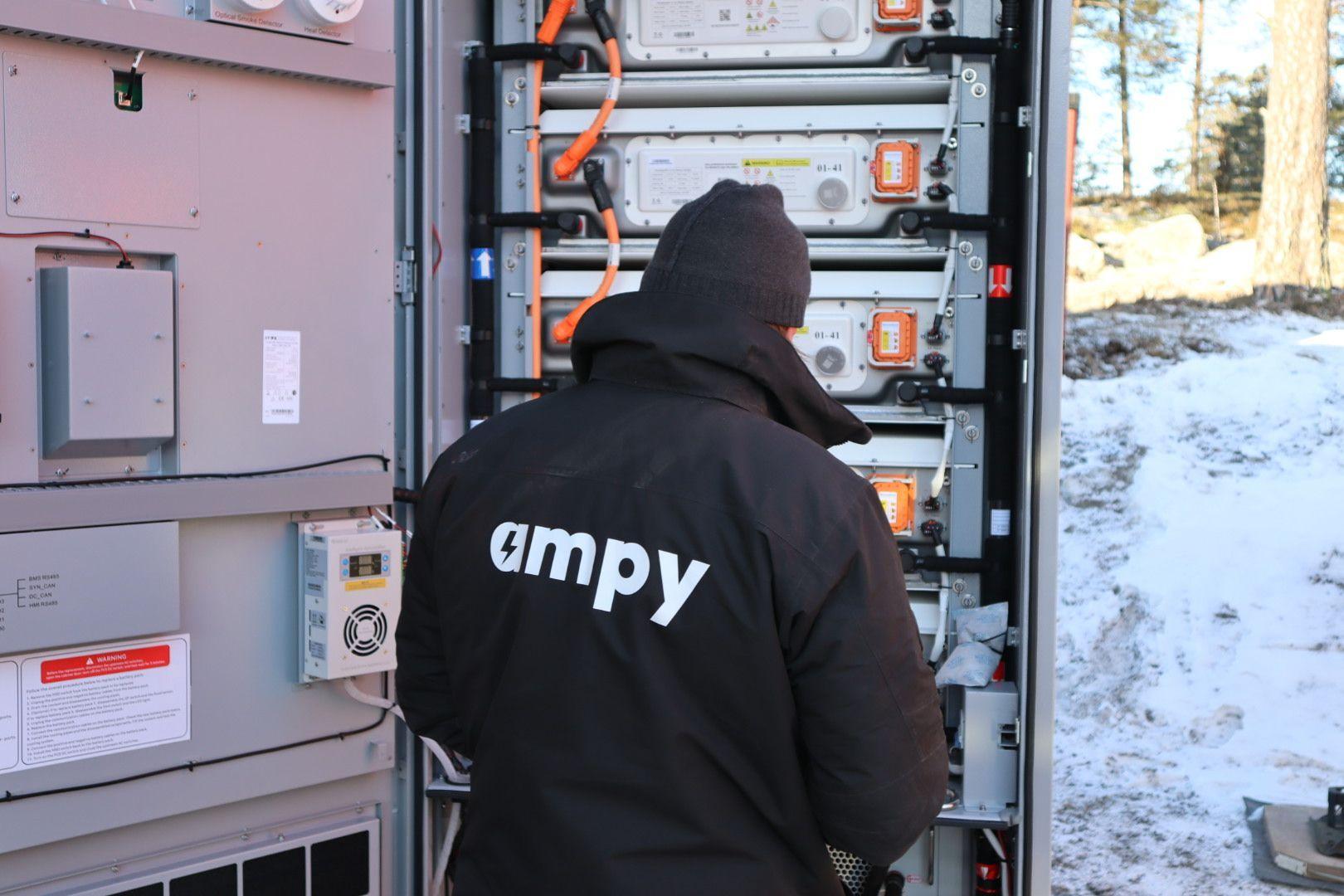 En tekniker i en svart jacka med logotypen "ampy" arbetar med batterier i en container i snöigt landskap.