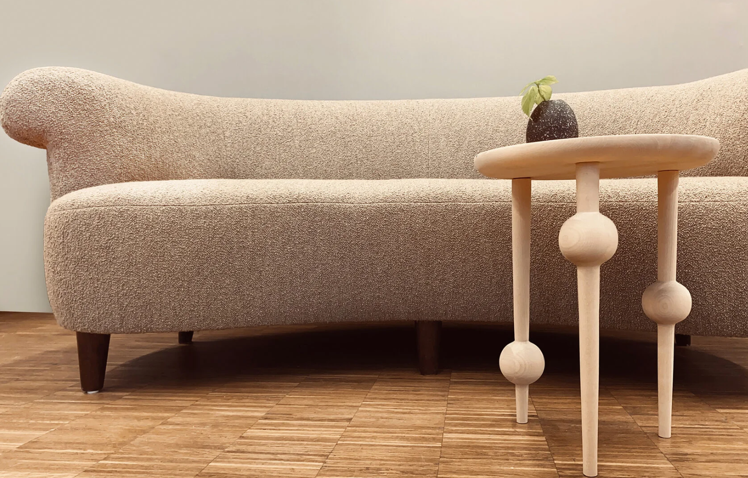 En modern soffa i beige tyg med rundade kanter, bredvid ett litet bord med tre ben och en växt i en kruka.
