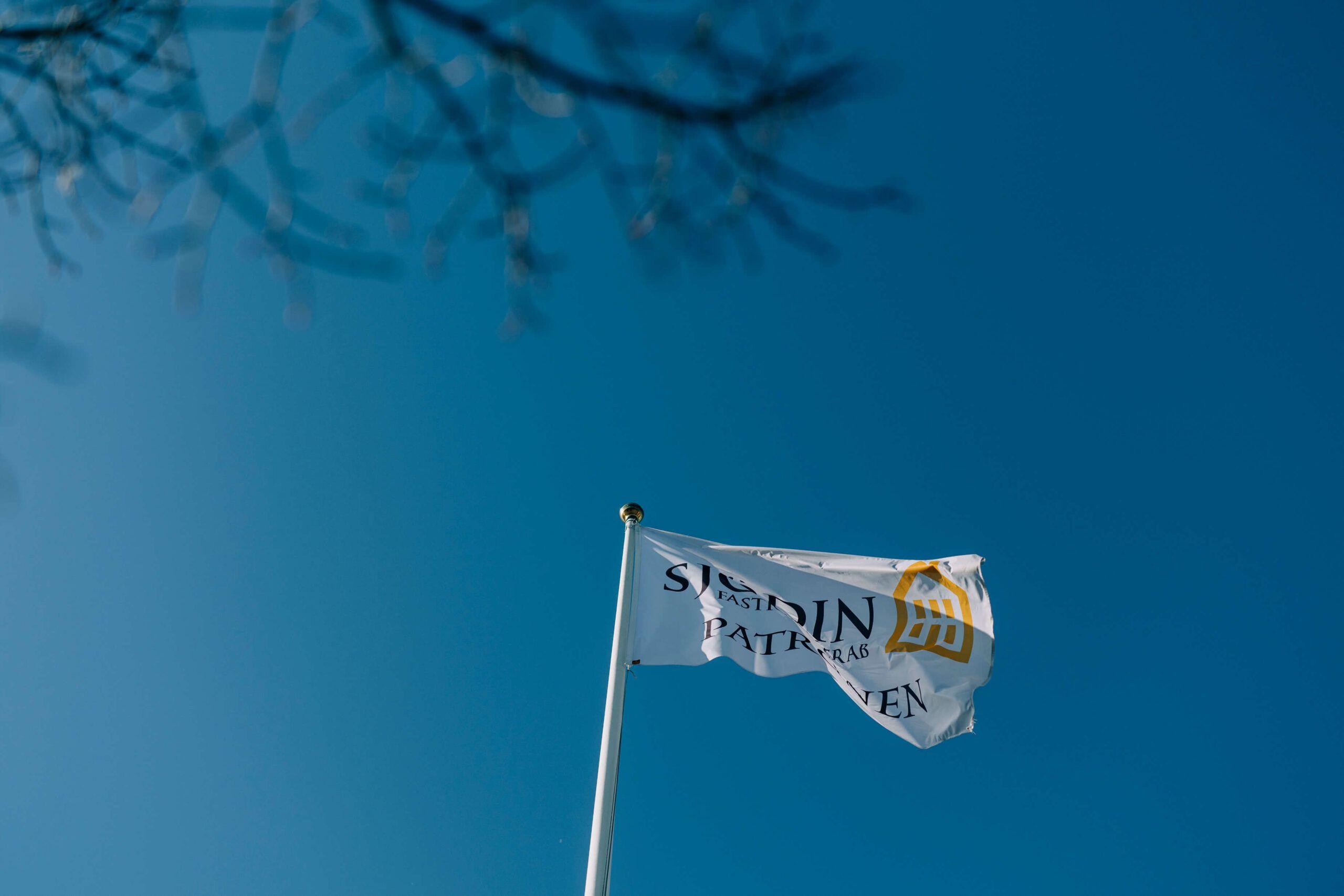 En vit flagga med texten "Svensin" vajar i vinden mot en klarblå himmel.