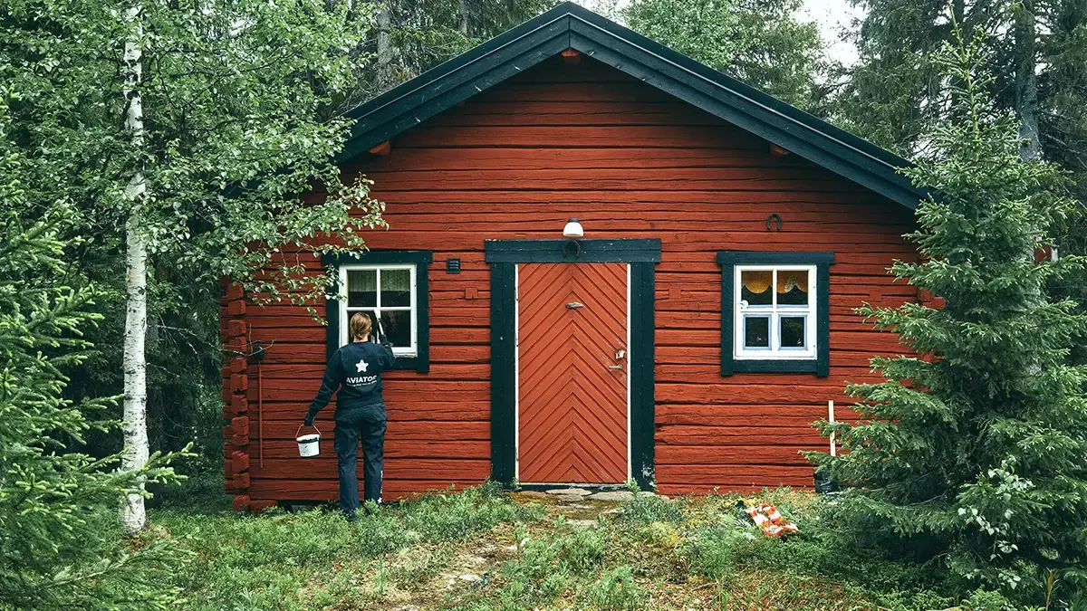 En person som håller en hink står framför ett rött hus omgivet av gräs och träd.