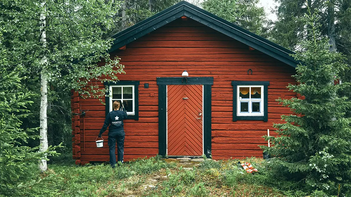 En person som håller en hink står framför ett rött hus omgivet av gräs och träd.